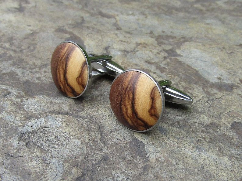 May include: A pair of cufflinks with a round, wooden design. The wood is a light brown with darker brown streaks. The cufflinks are made of silver metal.