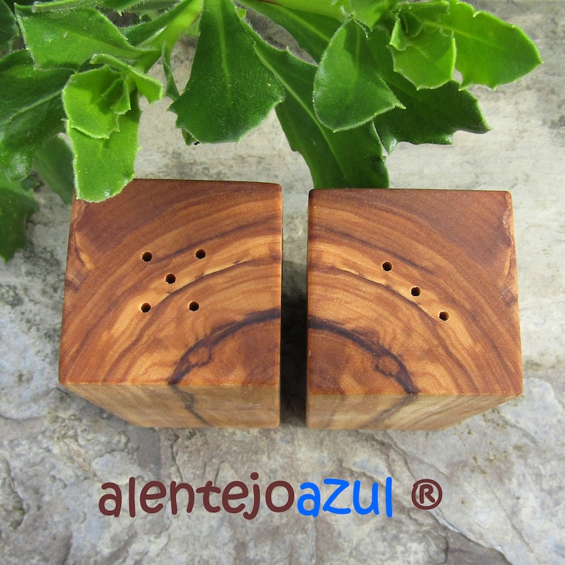 Op de afbeelding: Een set van twee houten zout- en peperstrooiers gemaakt van olijfhout. De strooiers zijn vierkant en hebben een natuurlijke houtkleur. Elke strooier heeft vijf gaten aan de bovenkant om zout en peper te strooien.