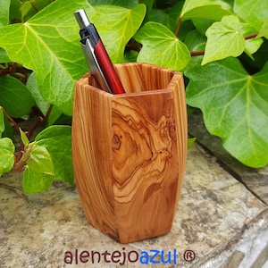 Pode incluir: Um porta-canetas de madeira feito de madeira de oliveira. O porta-canetas tem uma forma hexagonal e é de cor castanho natural com um padrão de grão de madeira. Uma caneta prateada e vermelha está dentro do porta-canetas.
