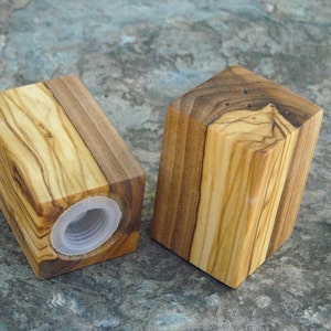 May include: Two wooden salt and pepper shakers made from olive wood. The shakers are square in shape and have a natural wood finish. The shakers have a small hole on the top for dispensing salt and pepper.