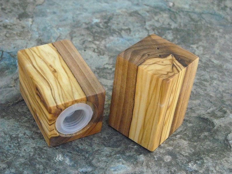 May include: Two wooden salt and pepper shakers made from olive wood. The shakers are square in shape and have a natural wood finish. The shakers have a small hole on the top for dispensing salt and pepper.