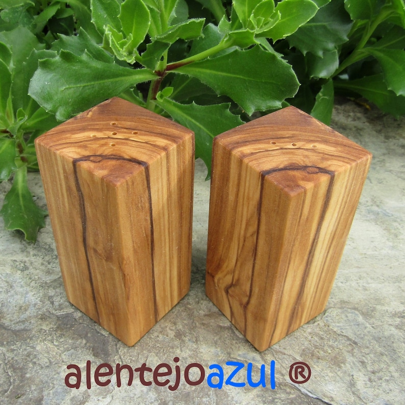 Op de afbeelding: Twee houten zout- en peperstrooiers gemaakt van olijfhout. De strooiers zijn vierkant en hebben een natuurlijke houtkleur. De strooiers staan op een grijs oppervlak.