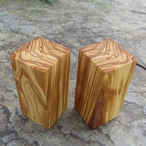 Op de afbeelding: Twee houten zout- en peperstrooiers gemaakt van olijfhout. De strooiers zijn vierkant en hebben een natuurlijke houten afwerking. Ze hebben gaten aan de bovenkant om zout en peper te strooien.