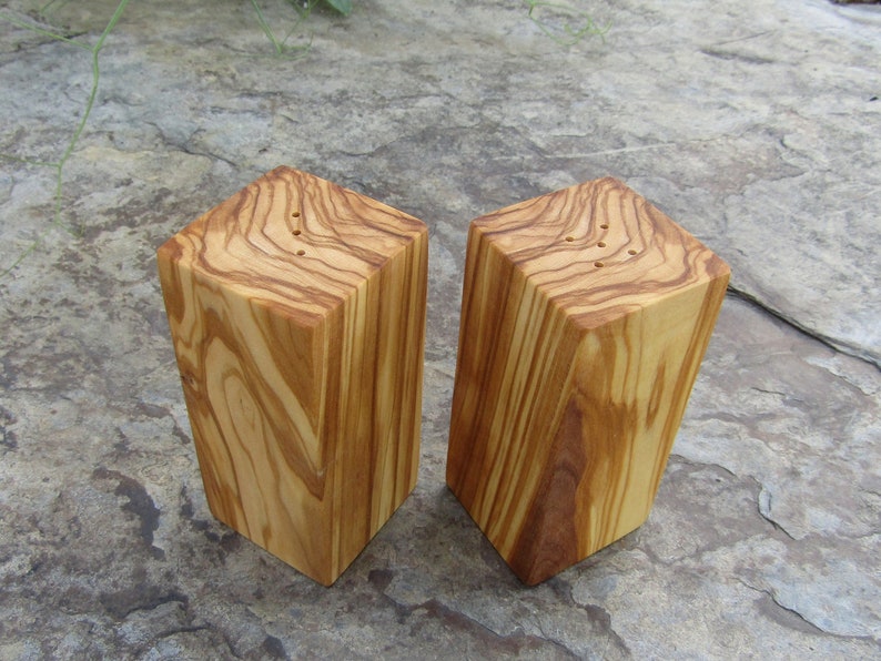 Op de afbeelding: Twee houten zout- en peperstrooiers gemaakt van olijfhout. De strooiers zijn vierkant en hebben een natuurlijke houten afwerking. Ze hebben gaten aan de bovenkant om zout en peper te strooien.