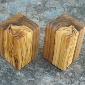May include: Two square wooden salt and pepper shakers made from olive wood. The shakers have a natural wood finish and are approximately 2 inches tall.