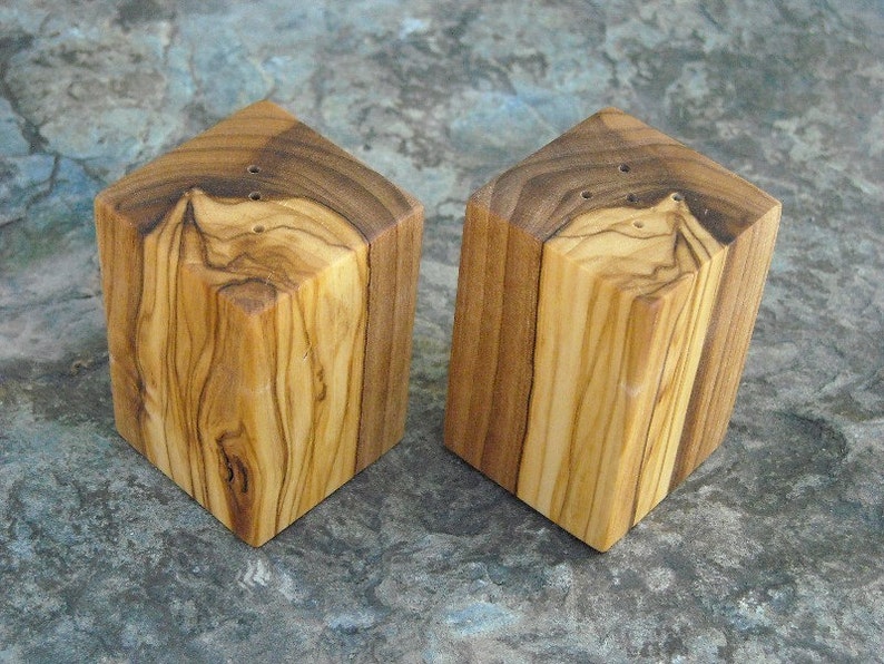 May include: Two square wooden salt and pepper shakers made from olive wood. The shakers have a natural wood finish and are approximately 2 inches tall.
