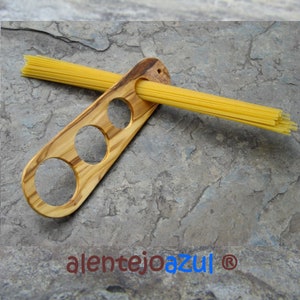 Könnte beinhalten: Ein Spaghetti-Halter aus Holz mit drei unterschiedlich großen Löchern zum Aufbewahren von Spaghetti. Der Halter ist aus Olivenholz gefertigt und hat eine natürliche Oberfläche. Ein Bündel Spaghetti liegt auf dem Halter.