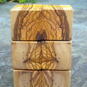 May include: A set of four wooden salt and pepper shakers made from olive wood. The shakers are stacked on top of each other and have a natural wood finish. Each shaker has a series of small holes drilled into the top for dispensing salt or pepper.