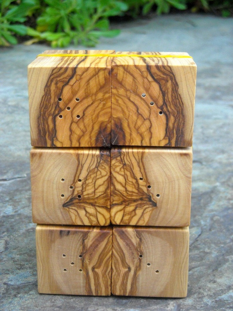 May include: A set of four wooden salt and pepper shakers made from olive wood. The shakers are stacked on top of each other and have a natural wood finish. Each shaker has a series of small holes drilled into the top for dispensing salt or pepper.