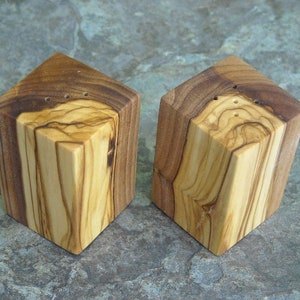 May include: Two square wooden salt and pepper shakers made from olive wood. The shakers have a natural wood grain pattern and are a light brown color.