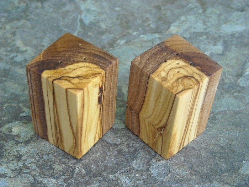 May include: Two square wooden salt and pepper shakers made from olive wood. The shakers have a natural wood grain pattern and are a light brown color.