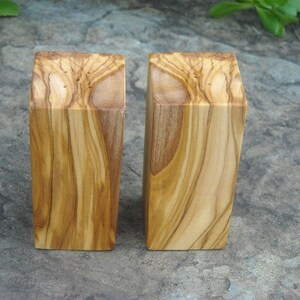 Op de afbeelding: Twee houten zout- en peperstrooiers gemaakt van olijfhout. De strooiers zijn rechthoekig en hebben een natuurlijke houten afwerking.