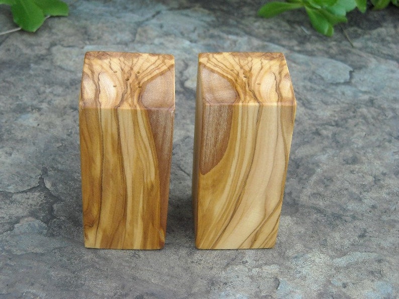 Op de afbeelding: Twee houten zout- en peperstrooiers gemaakt van olijfhout. De strooiers zijn rechthoekig en hebben een natuurlijke houten afwerking.
