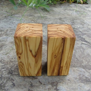 Op de afbeelding: Twee houten zout- en peperstrooiers gemaakt van olijfhout. De strooiers zijn rechthoekig en hebben een natuurlijke houtkleur. Ze hebben gaten aan de bovenkant om zout en peper te strooien.