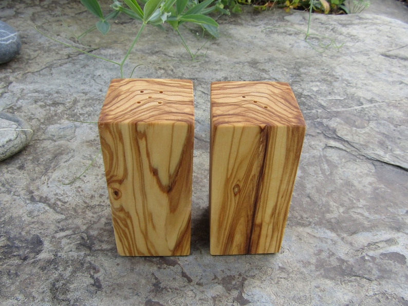 Op de afbeelding: Twee houten zout- en peperstrooiers gemaakt van olijfhout. De strooiers zijn rechthoekig en hebben een natuurlijke houtkleur. Ze hebben gaten aan de bovenkant om zout en peper te strooien.