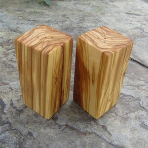 Op de afbeelding: Een set van twee houten zout- en peperstrooiers gemaakt van olijfhout. De strooiers zijn rechthoekig en hebben een natuurlijke houtkleur. Ze hebben kleine gaatjes bovenaan om zout en peper te strooien.