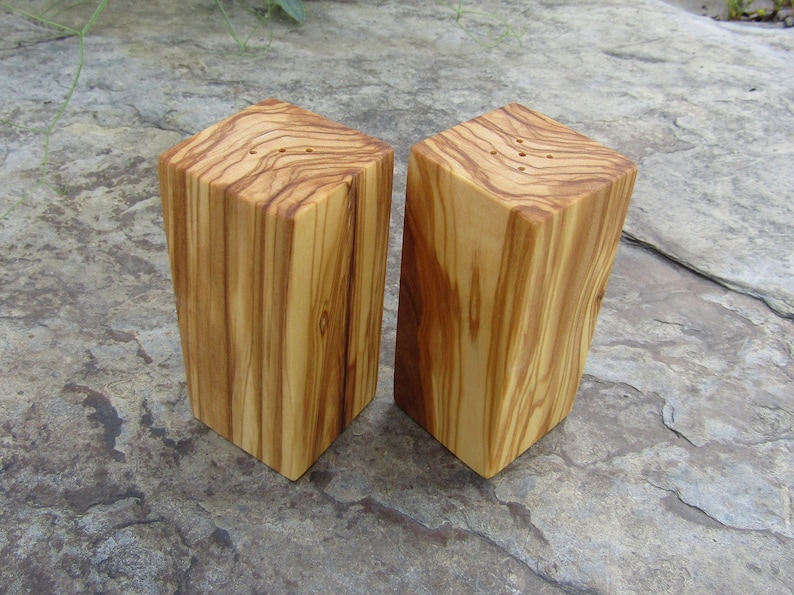 Op de afbeelding: Een set van twee houten zout- en peperstrooiers gemaakt van olijfhout. De strooiers zijn rechthoekig en hebben een natuurlijke houtkleur. Ze hebben kleine gaatjes bovenaan om zout en peper te strooien.