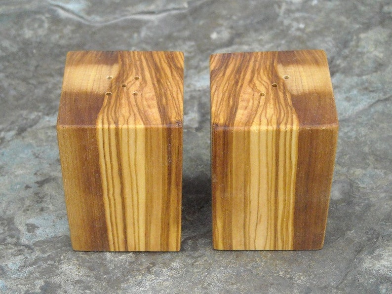 May include: A set of two wooden salt and pepper shakers with a natural wood grain finish. The shakers are square in shape and have a small hole on the top for dispensing salt and pepper.
