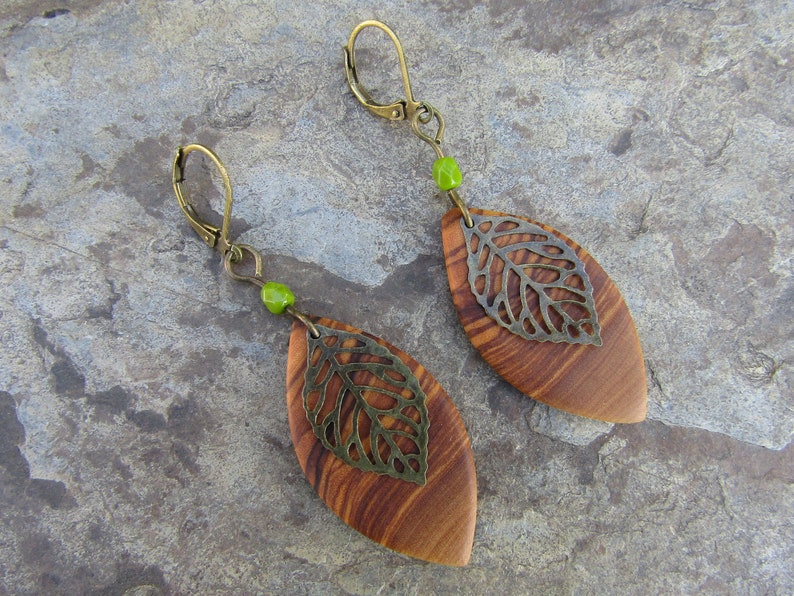 May include: A pair of wooden leaf earrings with a metal leaf design and green beads. The earrings are brown and have a natural wood grain.
