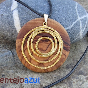 Pode incluir: Um pingente redondo de madeira com um design de círculos concêntricos de metal dourado. O pingente está pendurado em um cordão de couro preto.