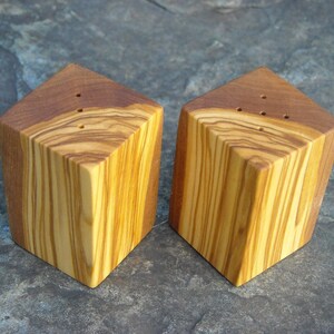 May include: Two wooden salt and pepper shakers made from olive wood. The shakers are square with rounded edges and have a natural wood grain pattern. The shakers have small holes on the top for dispensing salt and pepper.