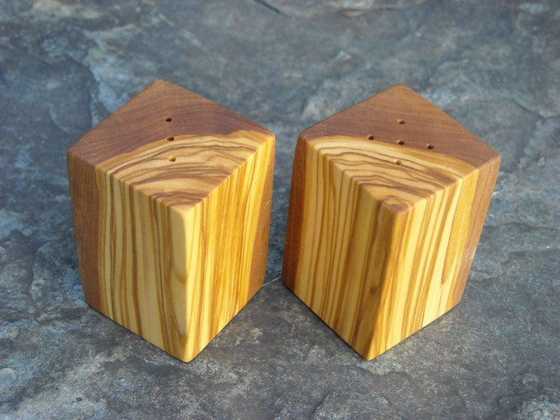 May include: Two wooden salt and pepper shakers made from olive wood. The shakers are square with rounded edges and have a natural wood grain pattern. The shakers have small holes on the top for dispensing salt and pepper.