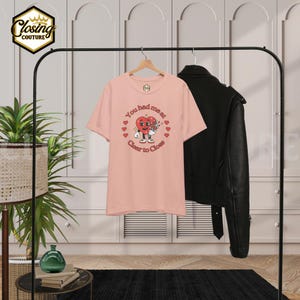 May include: A light pink t-shirt with the text "You had me at Clear to Close" and a cartoon heart design. The shirt is hanging on a black clothing rack with a black leather jacket.