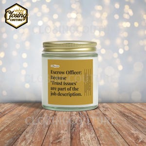 May include: A white candle in a glass jar with a gold lid. The label reads "Escrow Officer: Because 'Trust Issues' are part of the job description." The candle sits on a wooden surface.