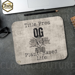 May include: A light beige mousepad with rounded corners featuring the text "Title Pros the OG Plant-Based Life" in black. The design includes a tree-like graphic made of stacked boxes. A laptop, pencil, and glasses are in the background.