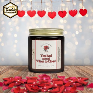 May include: A brown glass jar candle with a gold lid and a label that reads "You had me at 'Clear to Close'". The candle is decorated with red hearts and rose petals, perfect for Valentine's Day.