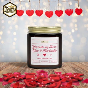 May include: A dark glass jar candle with a gold lid and a white label that reads "You make my Heart Clear & Marketable" with a heart and arrow graphic. Red heart decorations hang above the candle, and red rose petals are scattered below.