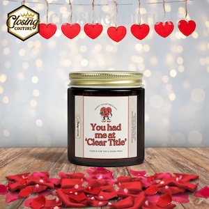 May include: A Valentine's Day candle in a glass jar with a gold lid. The label reads "You had me at 'Clear Title'" with a heart graphic. Red heart decorations hang above, and red rose petals are scattered below.