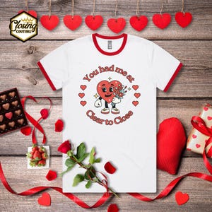 May include: White t-shirt with red trim featuring a heart character holding flowers, surrounded by hearts, and the text "You had me at Clear to Close." The shirt is on a wooden background with Valentine's Day decorations.