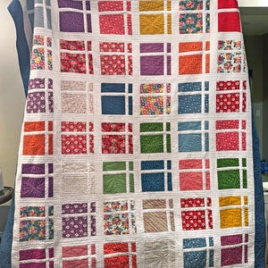 May include: A colorful quilt with a grid pattern. Each square features a different floral print fabric. The quilt is made with white fabric and has a blue border.