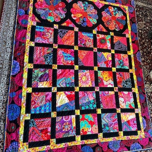 May include: A colorful quilt with a black background and a geometric pattern. The quilt is made up of squares and rectangles, and the fabric is a mix of bright colors, including red, pink, yellow, blue, and green.