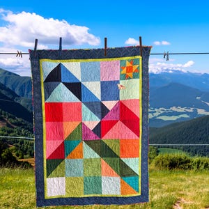 Può includere: Una colorata trapunta con motivi geometrici è appesa a un filo per il bucato sullo sfondo di un paesaggio montano. La trapunta presenta quadrati e triangoli nelle tonalità di rosso, rosa, blu, verde e arancione, con un bordo blu scuro e un bordo giallo e verde.