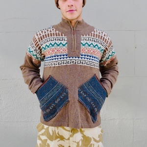 May include: A brown sweater with a colourful geometric pattern and blue knitted pockets. The sweater has a high neck and a zip closure. The person is wearing a brown and red patterned beanie.