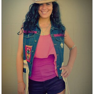 Op de afbeelding: Een vrouw draagt een denim vest met kleurrijke borduurwerk, een roze top en een donkerblauwe denim short. Ze draagt ook een breedgerande strohoed met een decoratieve band.