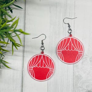 Op de afbeelding: Een paar rood-witte theatergordijn oorbellen. Elke oorbel heeft een cirkelvormig ontwerp met een rode achtergrond en een witte omtrek van een toneelgordijn. De oorbellen hangen aan zilverkleurige oorhaken.