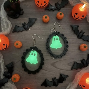 Op de afbeelding: Halloween-thema compositie met zwarte vleermuis- en pompoendecoraties, en twee paar zwart omlijste oorbellen met gloeiende groene spookontwerpen. Oranje pompoenkaarsen en een zwart kattenbeeldje zijn ook aanwezig.