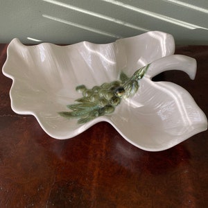 May include: A white ceramic leaf-shaped serving dish with a cluster of green grapes and leaves. The dish has a glossy finish and is placed on a dark wooden surface. Decorative tableware.