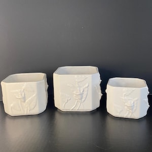 May include: A set of three white ceramic planters with an octagonal shape and embossed floral designs on the exterior. Each planter features a smooth, glossy finish and a consistent pattern of raised lily-like flowers and leaves on the sides.