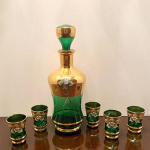 May include: A vintage green and gold decanter set. The decanter has a gold-colored body with green accents and a matching stopper. Six small glasses with floral designs and gold rims are arranged around the decanter.