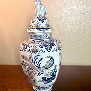 May include: A white and blue ceramic vase with a lid, featuring a cat figurine on top. The vase is decorated with a bird and floral design in blue, brown, and green. The vase has a hexagonal base.