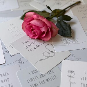 May include: A set of affirmation cards with a pink rose on top. The cards feature positive messages such as "I am allowed to feel stressed" and "I honor my limits and take time for myself".