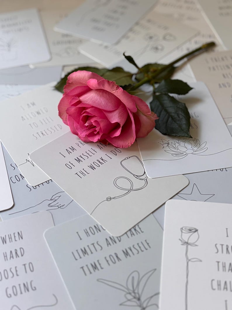 May include: A set of affirmation cards with a pink rose on top. The cards feature positive messages such as "I am allowed to feel stressed" and "I honor my limits and take time for myself".