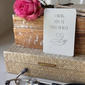 May include: A white card with the text "I bring hope to those in need" and a line drawing of a hand holding a heart. The card is sitting on a wooden box with a pink rose on top.