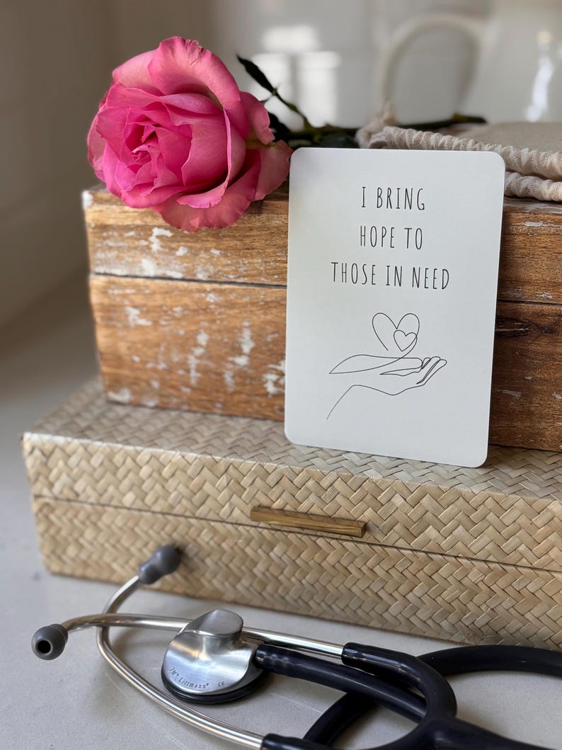 May include: A white card with the text "I bring hope to those in need" and a line drawing of a hand holding a heart. The card is sitting on a wooden box with a pink rose on top.