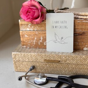 May include: A white card with the text "I have faith in my calling" and a simple line drawing of a dove with an olive branch in its beak. The card is sitting on top of a wooden box with a pink rose on top of the box. A stethoscope is lying on a woven surface.