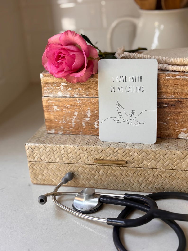 May include: A white card with the text "I have faith in my calling" and a simple line drawing of a dove with an olive branch in its beak. The card is sitting on top of a wooden box with a pink rose on top of the box. A stethoscope is lying on a woven surface.